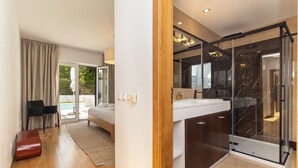 A modern bedroom with a bed, a desk, and a chair, a bathroom with a glass shower, and a view of a pool. - Villa da Oliveira with Pool - Golf Aroeira (Herdade da Aroeira)