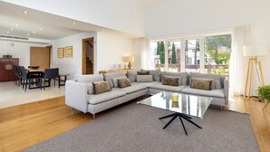 A modern living room with a sectional sofa, a glass coffee table, and a dining area with a wooden table and chairs. - Villa da Oliveira with Pool - Golf Aroeira (Herdade da Aroeira)
