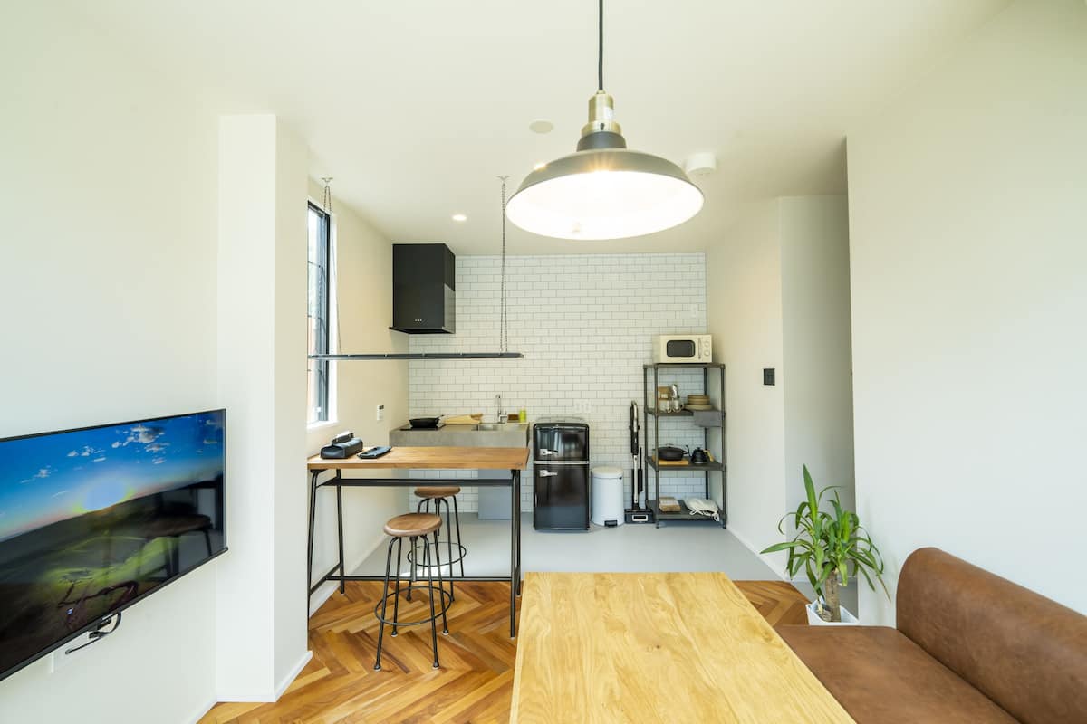 A modern kitchen with a wooden table, bar stools, a TV, and a hanging light fixture.