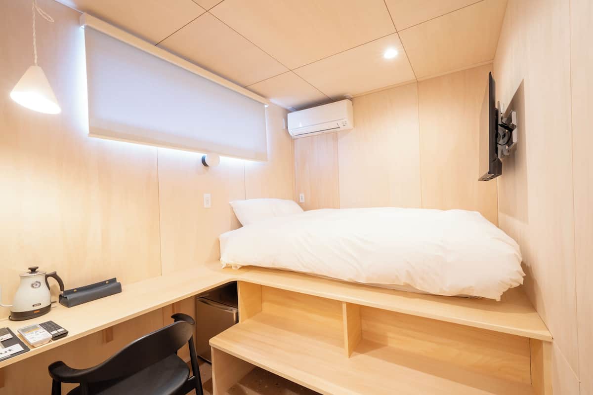 A compact room with a bed, a desk, a chair, a coffee maker, and an air conditioning unit.