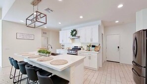 Une cuisine moderne avec des meubles blancs, un îlot central doté de tabourets, un réfrigérateur en acier inoxydable et un luminaire suspendu.