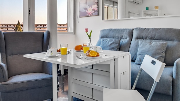 Et moderne værelse med et spisebord dækket til morgenmad, en grå sofa og et stort vindue med udsigt til bygninger.