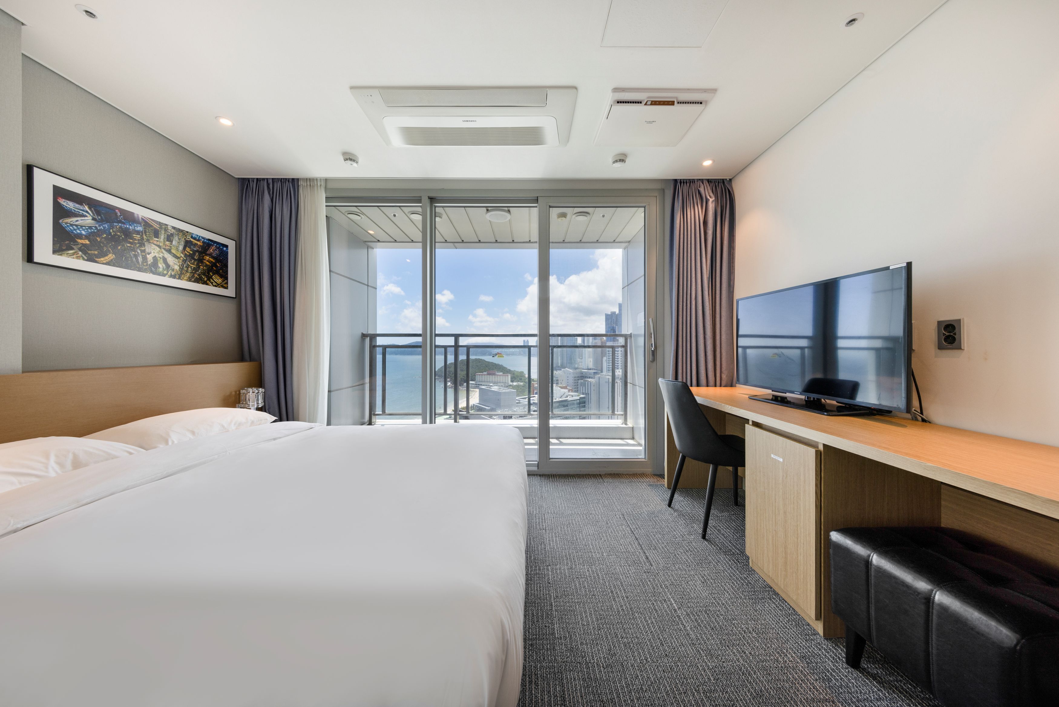 Habitación de hotel con cama, escritorio, televisión y vista de la ciudad a través de amplias ventanas.