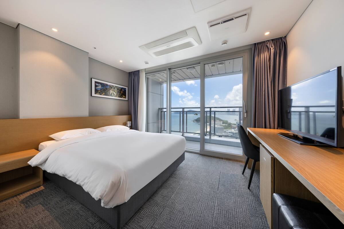 A hotel room with a large bed, a desk with a TV, a window with a view of the city, and a ceiling with recessed lighting.