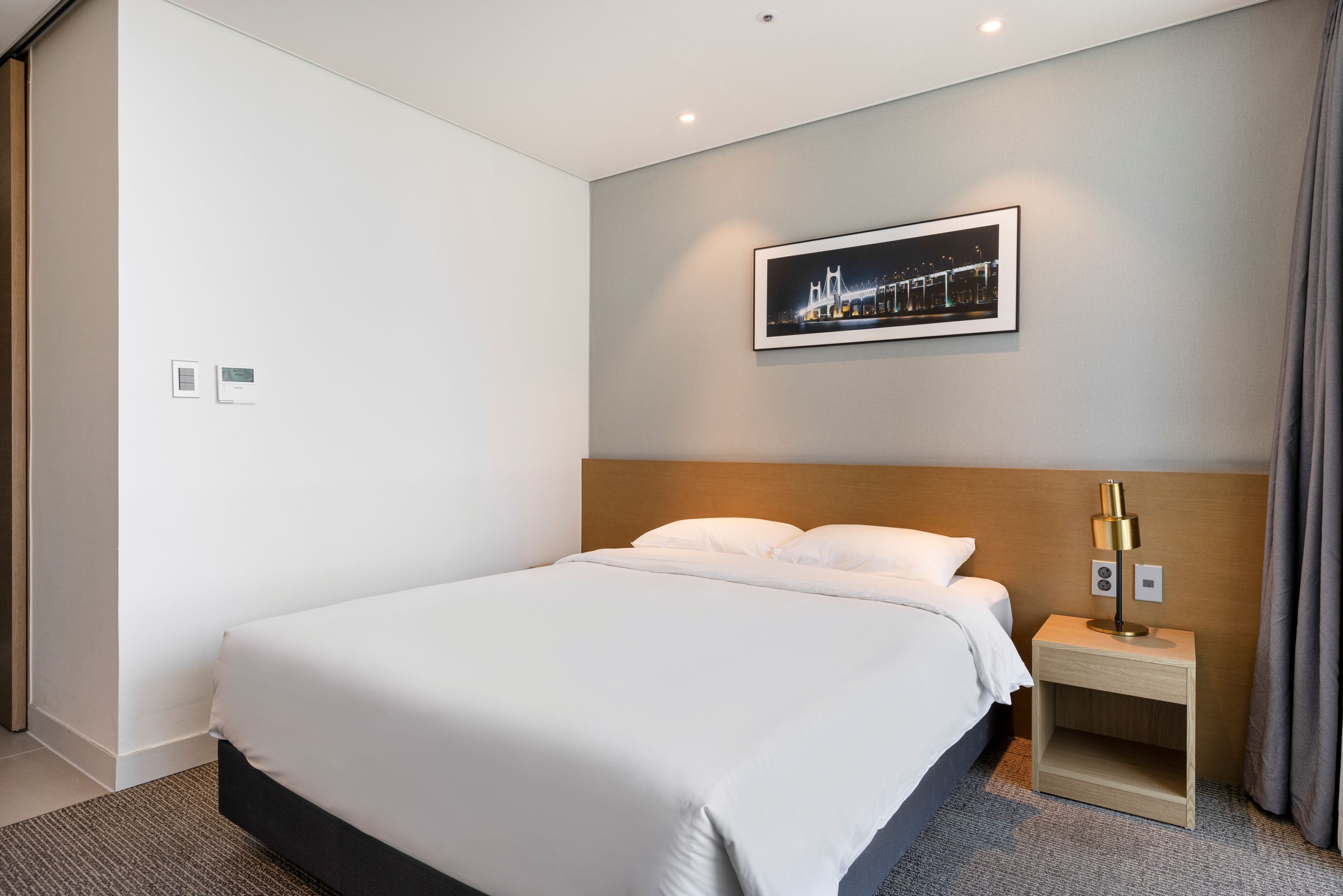 A modern hotel room with a large bed, a nightstand with a lamp, a framed picture of a bridge, and a control panel on the wall.