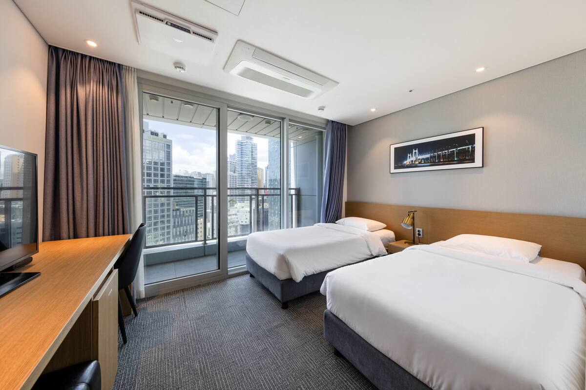 A hotel room with two beds, a desk, a chair, a TV, and a large window with a city view.