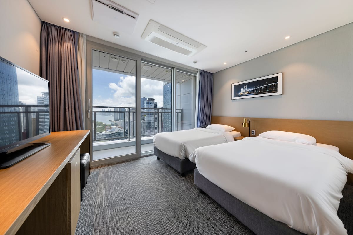 A hotel room with two beds, a desk, a TV, and a large window with a city view.