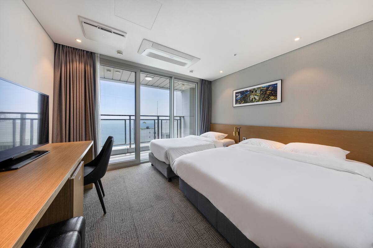 A hotel room with a desk, two beds, a large window with curtains, a painting on the wall, and a view of the city.