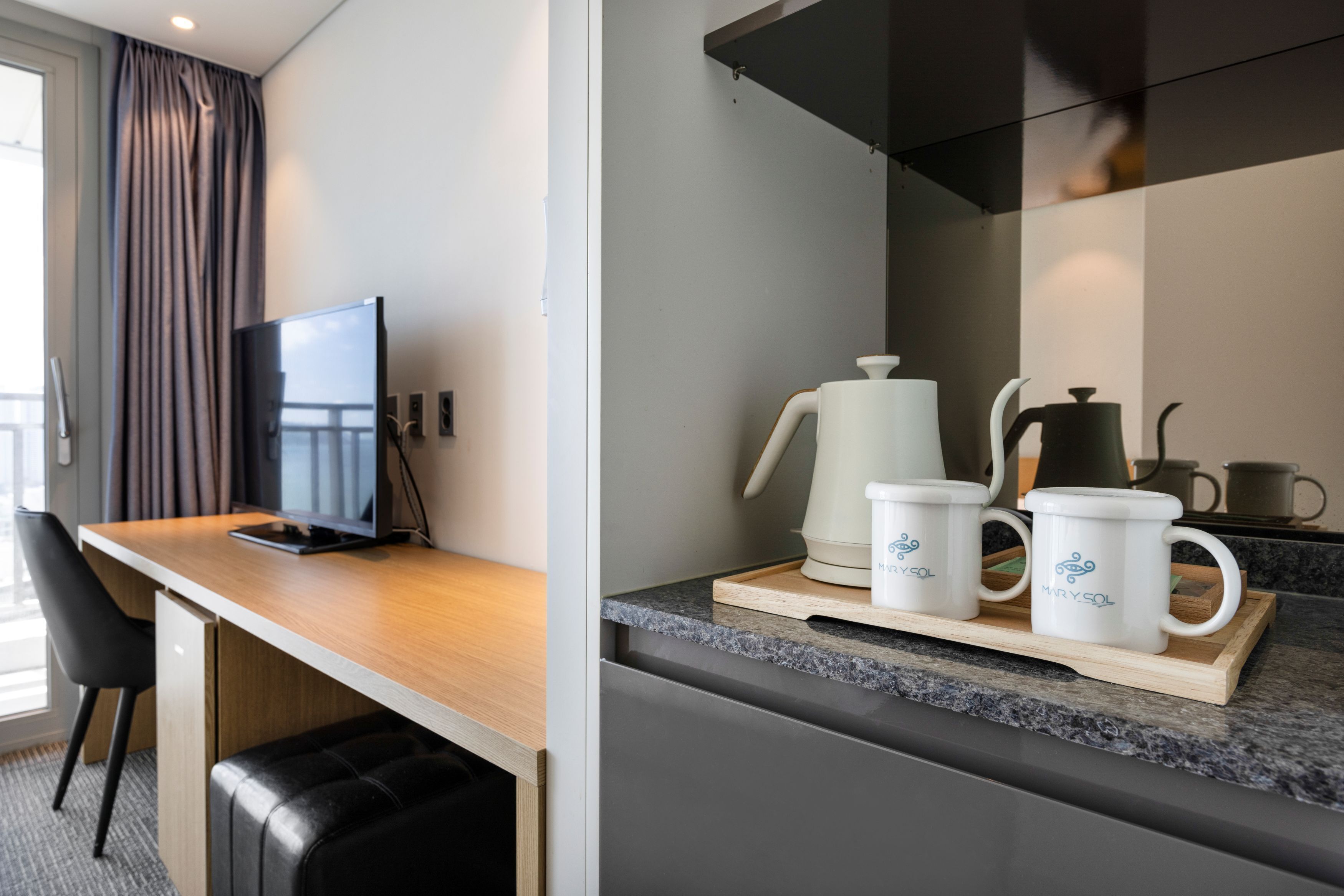 A modern hotel room with a wooden desk, a flat-screen TV, a comfortable black leather chair, a kitchen area with a coffee maker, two mugs, and a tray.