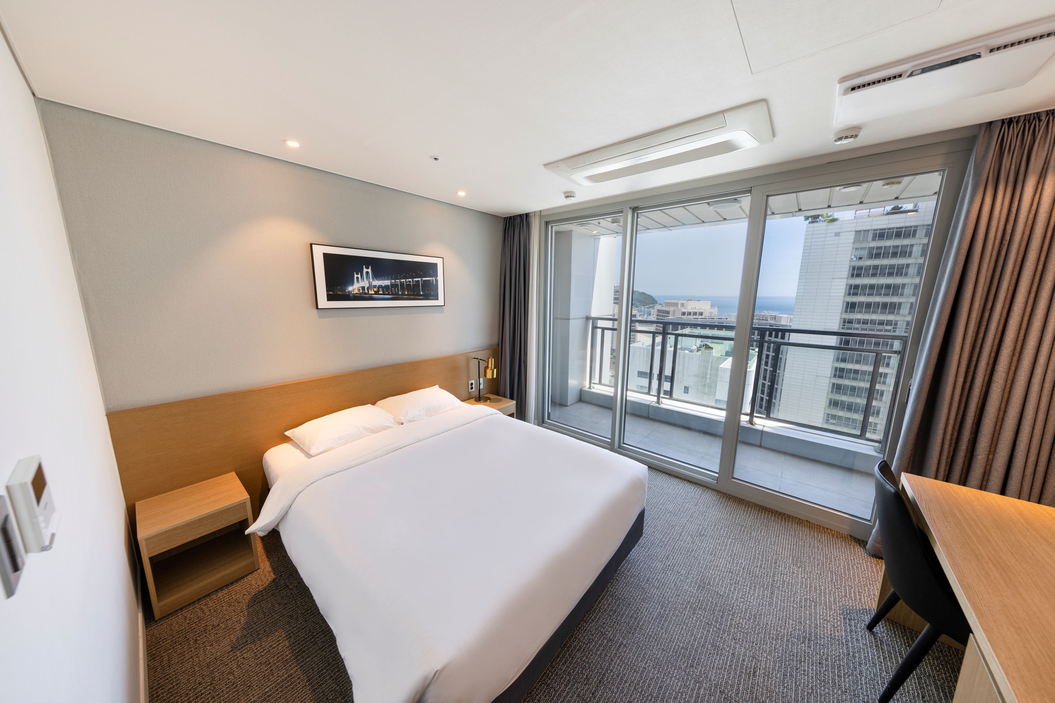 Habitación de hotel con una cama grande, un escritorio, una mesita pequeña, una mesita de noche, un cuadro en la pared y un balcón con vistas a la ciudad.