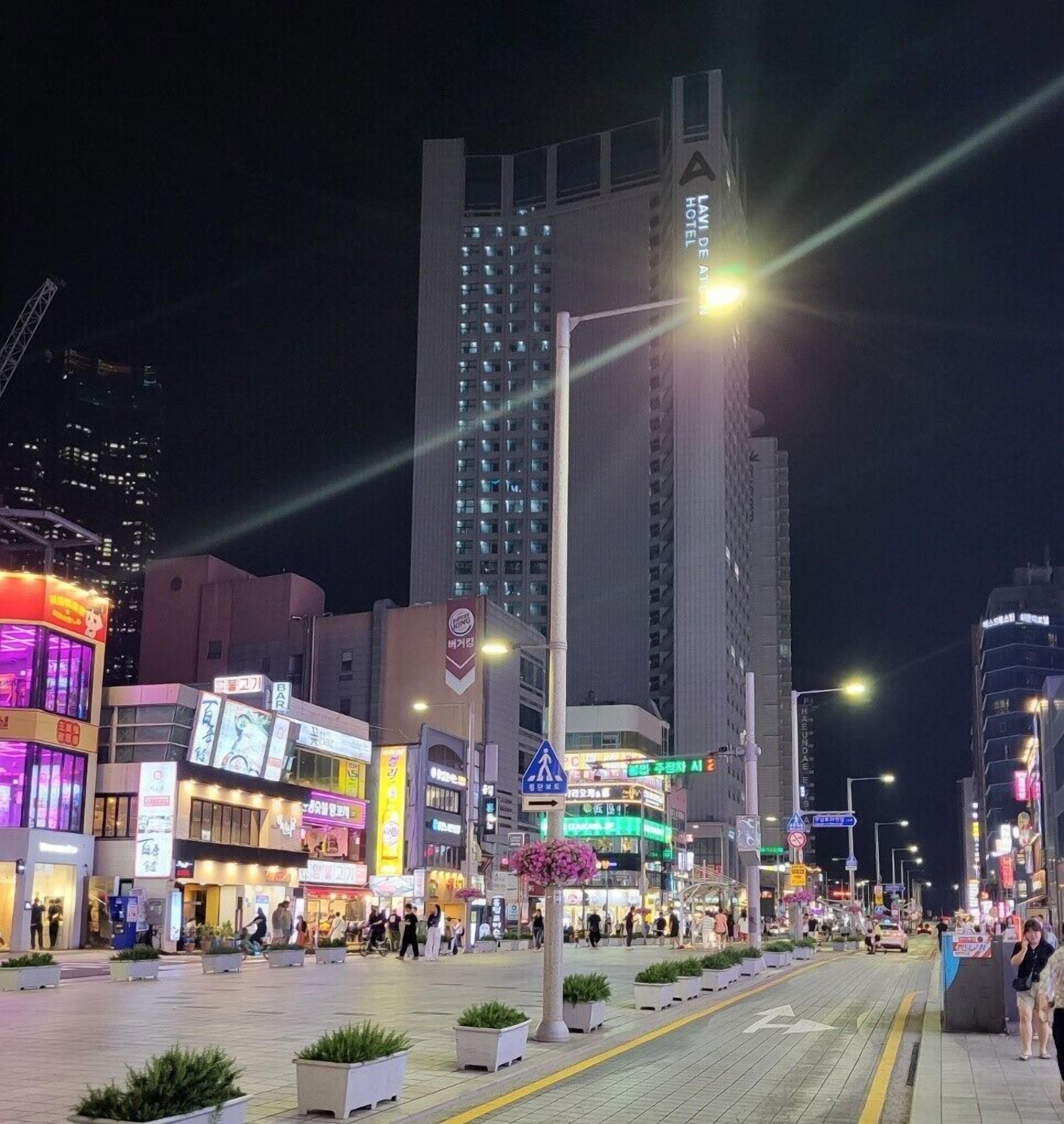A nighttime cityscape with a prominent hotel building, illuminated signage, and a pedestrian area with planters.