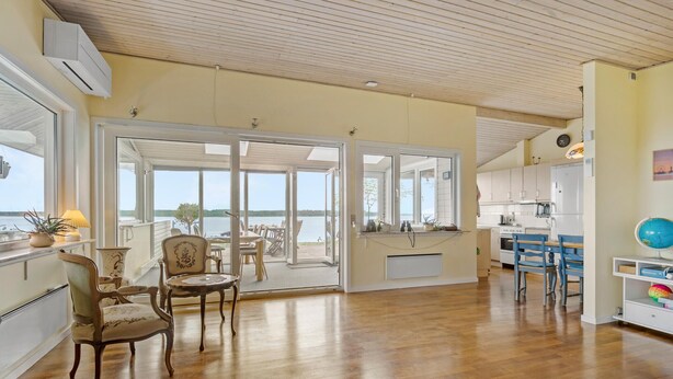 A spacious living room with a wooden floor, a dining area with a table and chairs, a kitchen with white cabinets, and a large sliding glass door leading to a balcony with a view of the water. - Gorgeous home in ølsted with kitchen (Ølsted)