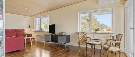 A spacious living area with wooden flooring, a red sofa, a TV, and two windows providing natural light.
