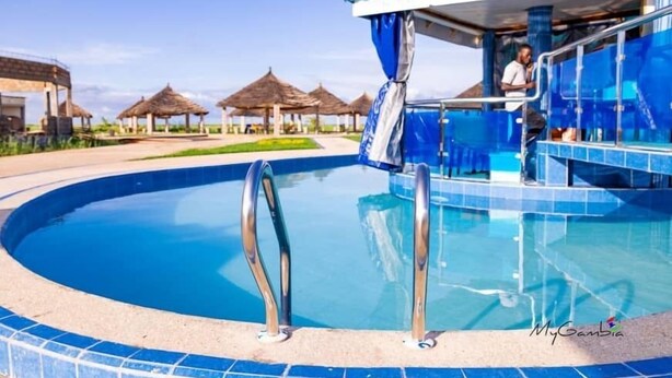 Une piscine dont le bord est recouvert de carreaux bleus, un espace abrité où se trouve une personne, et des constructions à toits de chaume en arrière-plan.