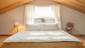 Ett sovrum med ett sluttande tak, en säng med vita sängkläder, ett sängbord med en lampa och ett fönster med gardiner.