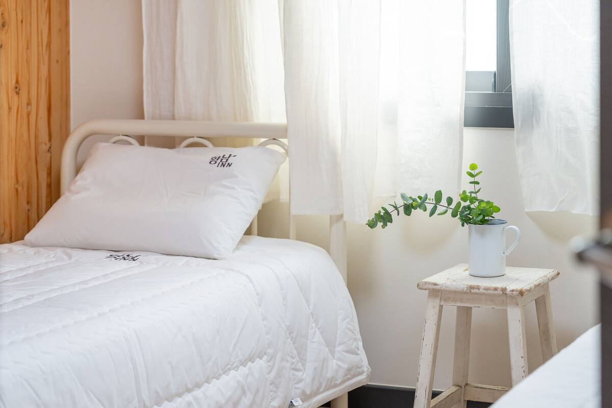 Ein Bett mit weißer Bettwäsche und Kissen, ein kleiner Nachttisch mit einer weißen Tasse und einer Pflanze, sowie durchsichtige Vorhänge an einem Fenster.