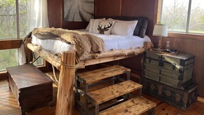 Quarto de cabana rústica com cama sobre plataforma de madeira, um baú de madeira e vista para árvores pela janela.