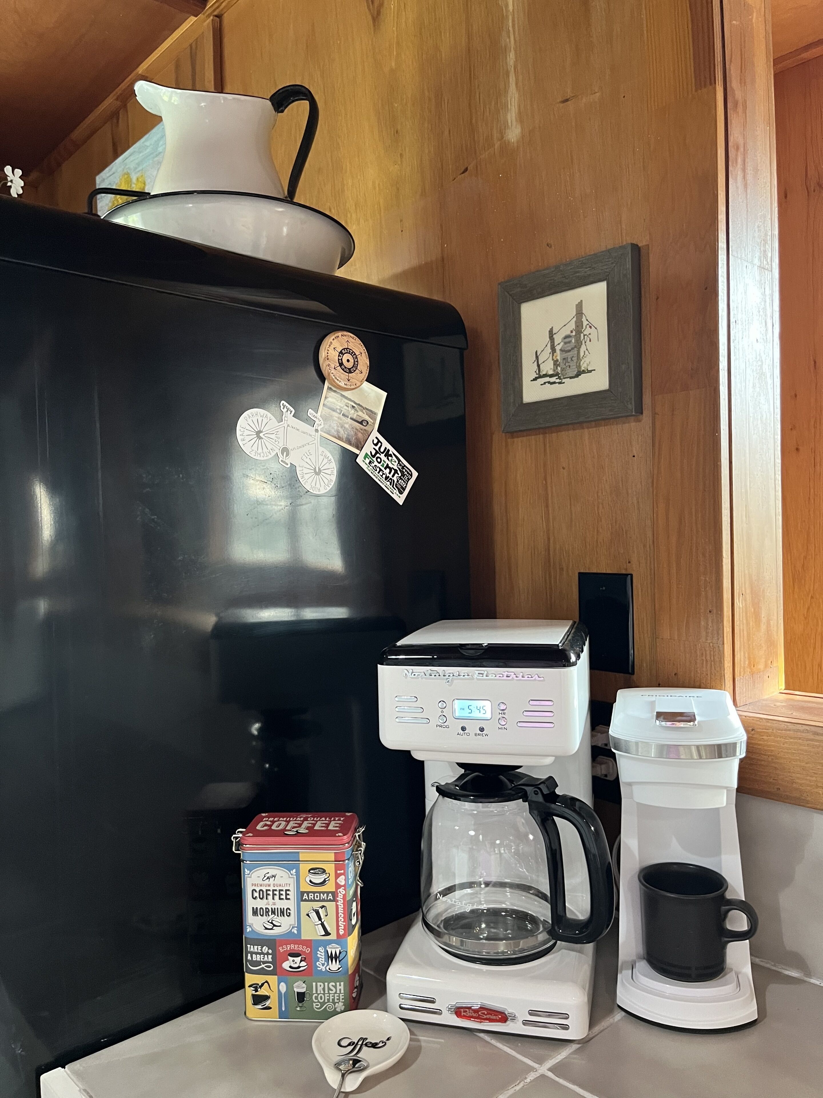 Een kitchenette met een koffiezetapparaat, een pak koffie, een mok en een koelkast.