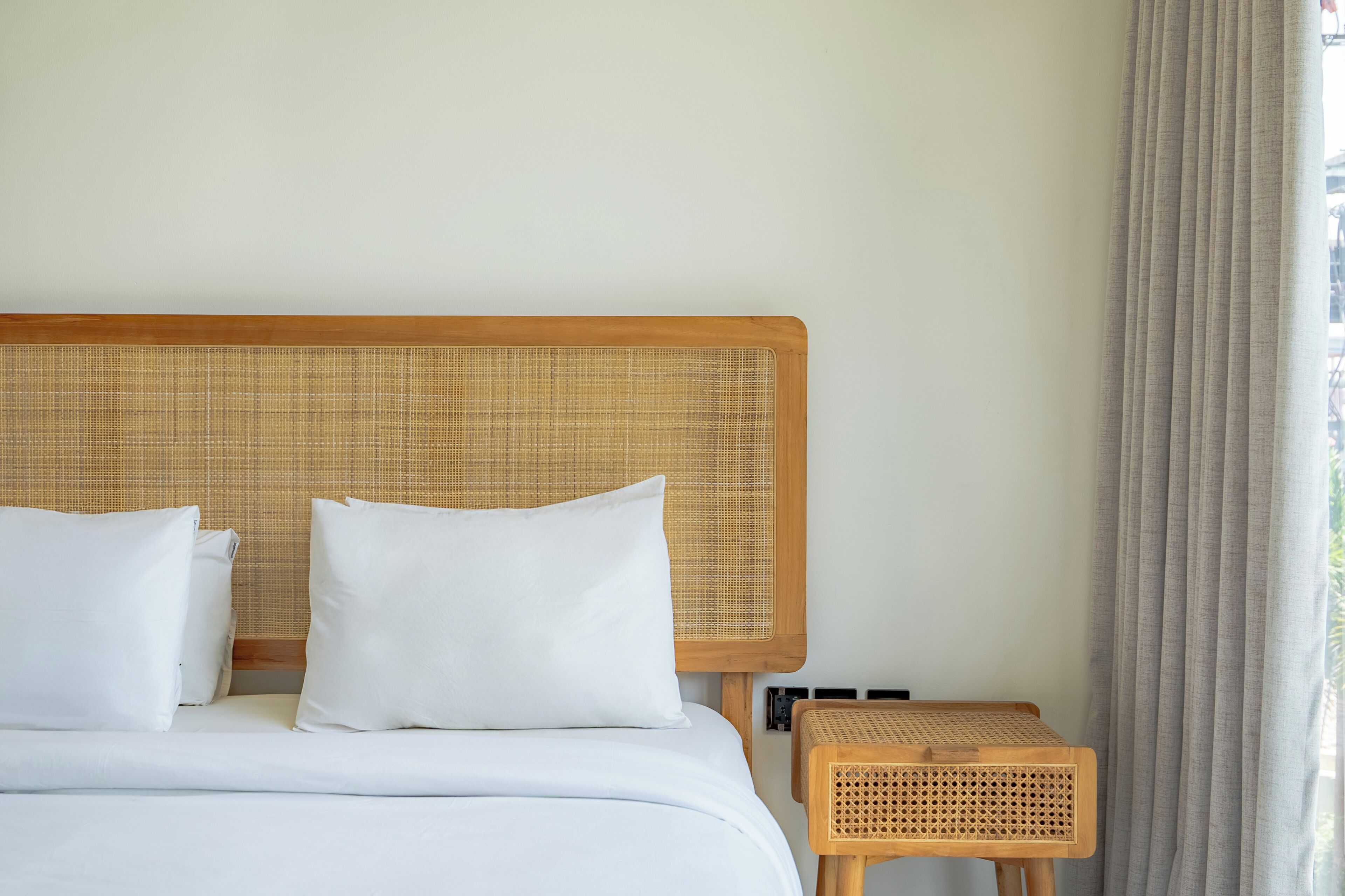 A hotel room with a bed, white bedding, a wooden headboard, a wicker nightstand, and a window with curtains.