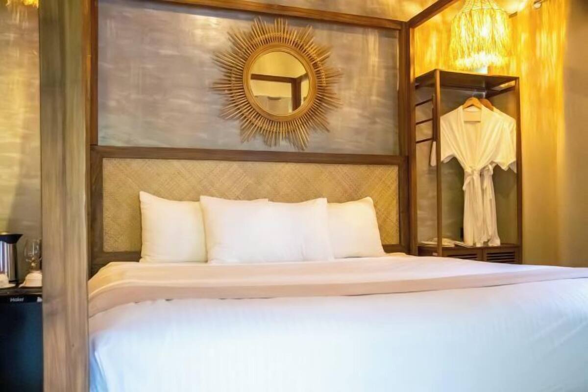 Una cama bien hecha con sábanas blancas, cabecera de madera, espejo con diseño de sol y una bata colgada.