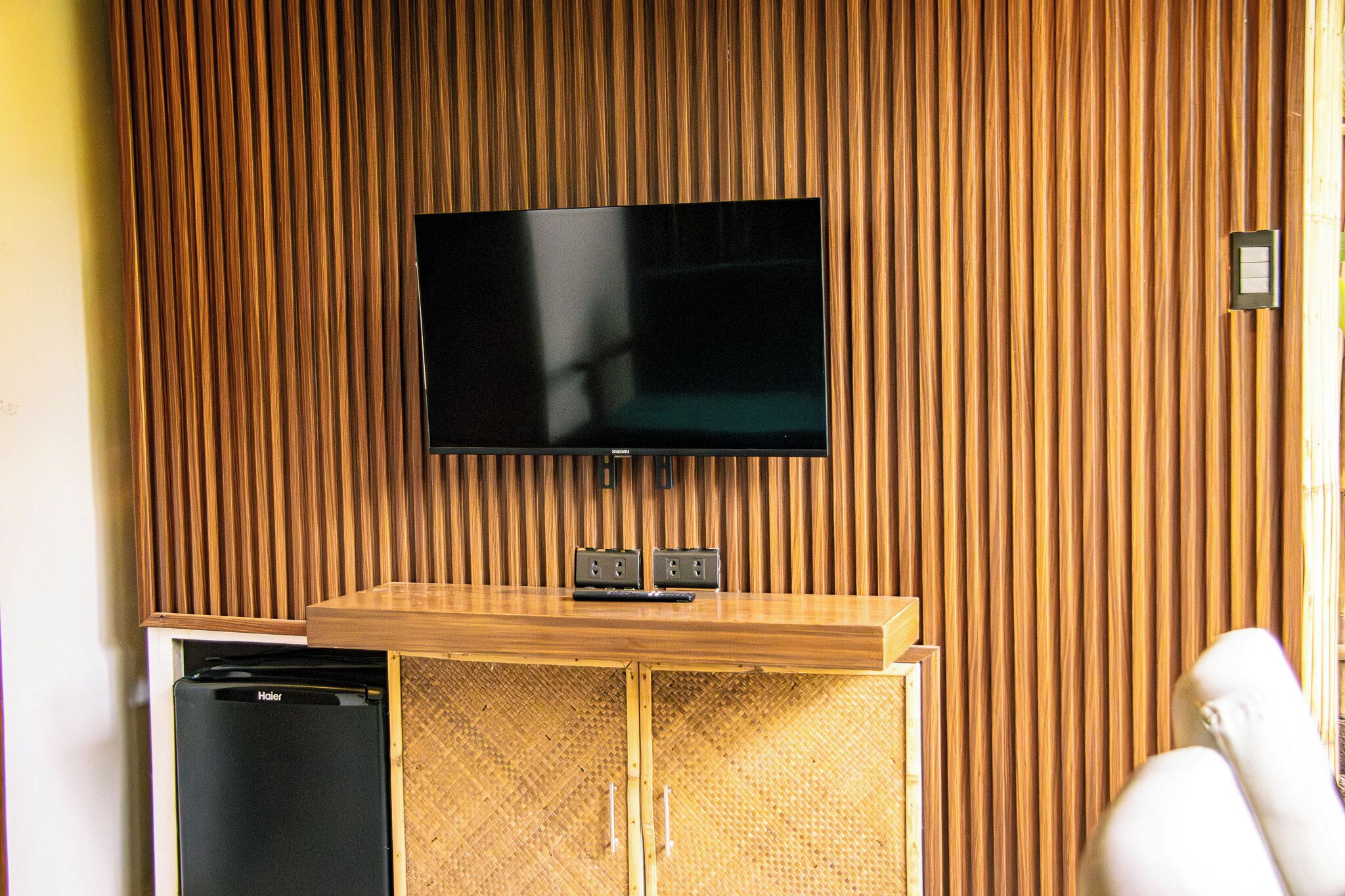 A room with a flat-screen TV mounted on a wooden wall, a wooden cabinet with two electrical outlets, and a small black appliance.