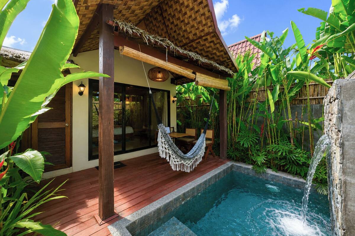 A pool with a hammock, wooden deck, and lush tropical plants.