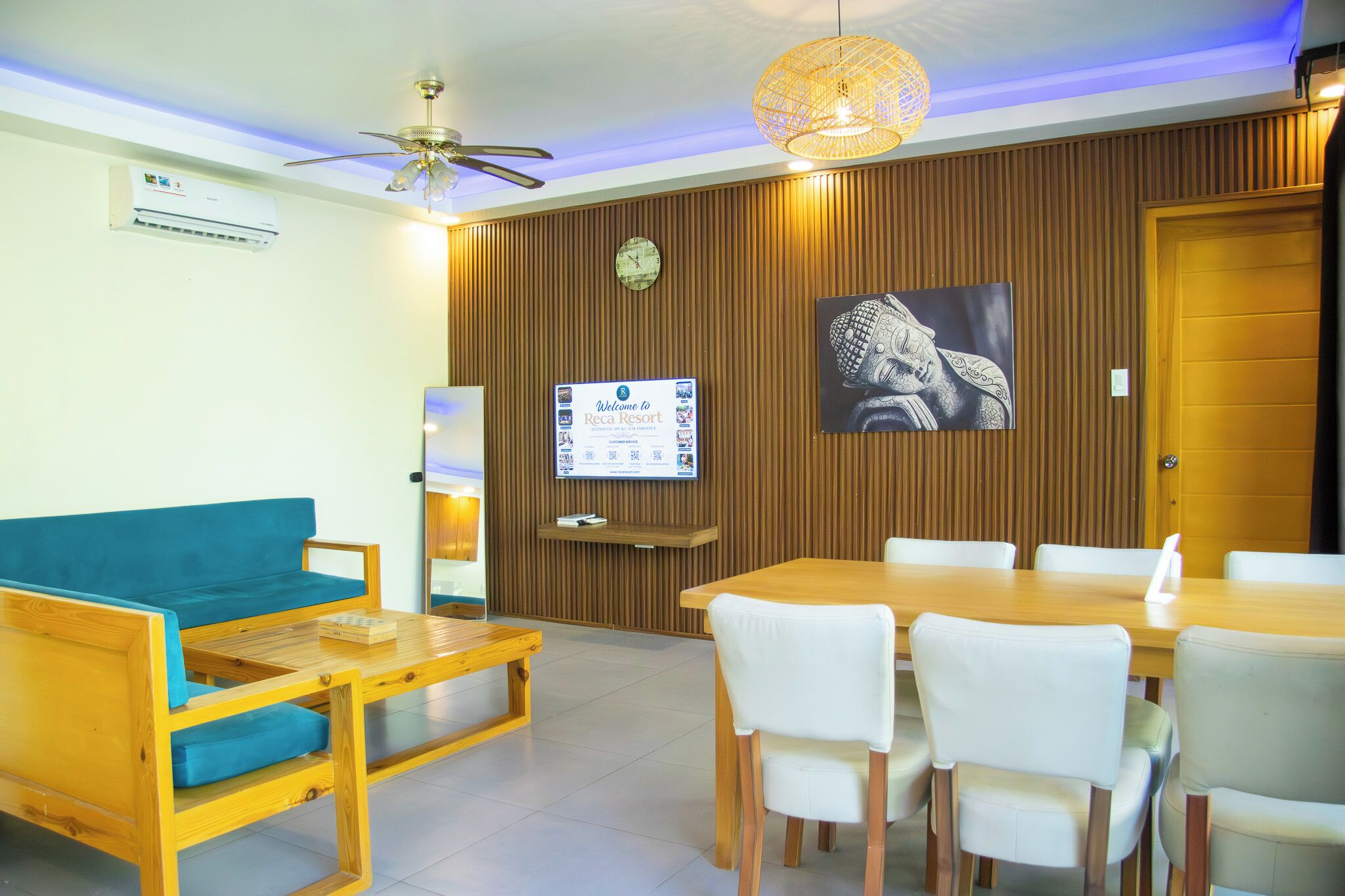 A hotel room with a wooden ceiling fan, a television, a clock, a framed artwork, a sofa, a dining table with chairs, and a small wooden table.