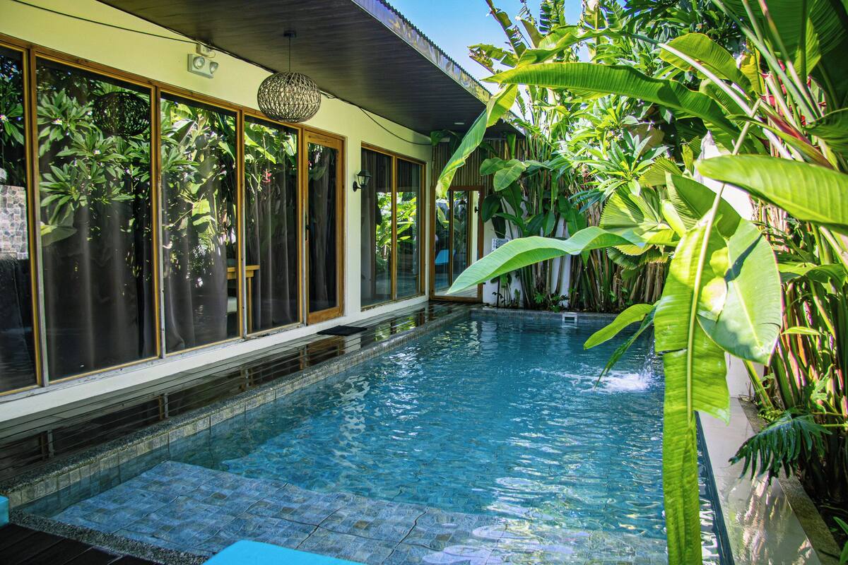 A swimming pool with clear water, surrounded by lush tropical plants and large windows.