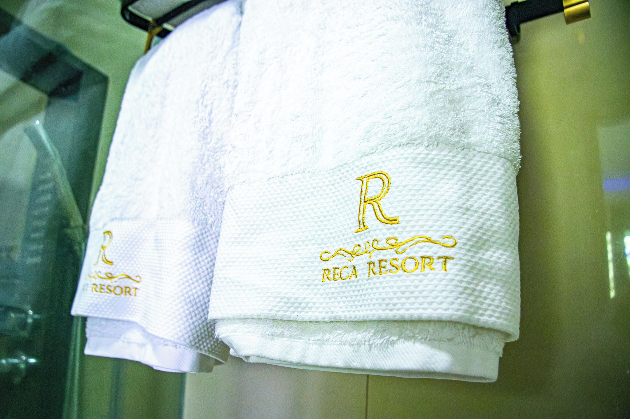 Two white towels with embroidered text, one reads 'RECA RESORT' and the other has a letter 'R' and the word 'RESORT'.