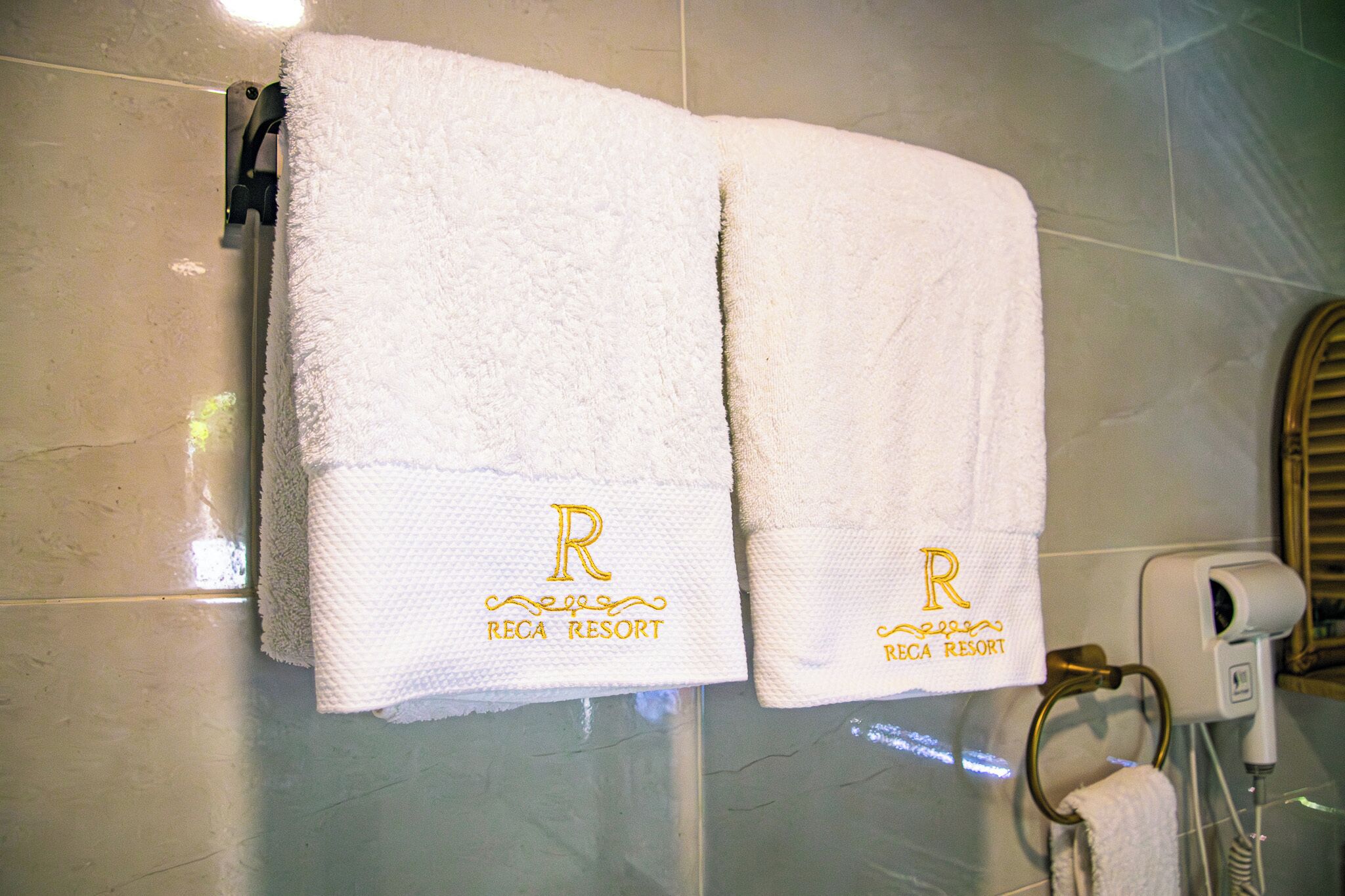 Two white towels with embroidered 'R' and 'RECA RESORT' hanging on a tiled wall.