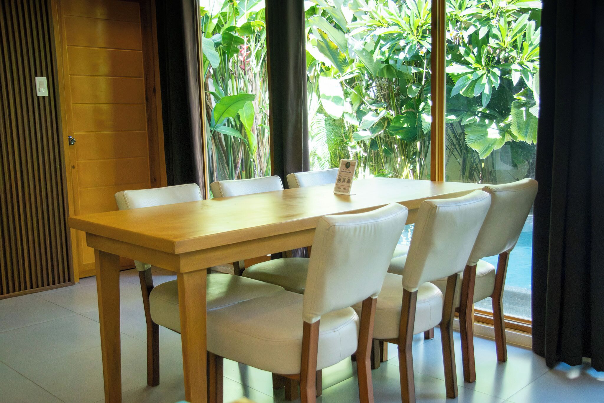 Un coin repas avec une table en bois et six chaises, de grandes fenêtres donnant sur un paysage verdoyant et une porte en bois.