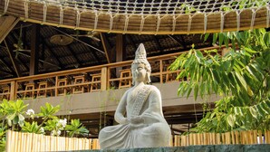A statue of a seated figure in a courtyard with a wooden fence, a thatched roof structure, and lush greenery. - RECA Resort Philippines | Luxury Villas & Nature Retreat (Porac)