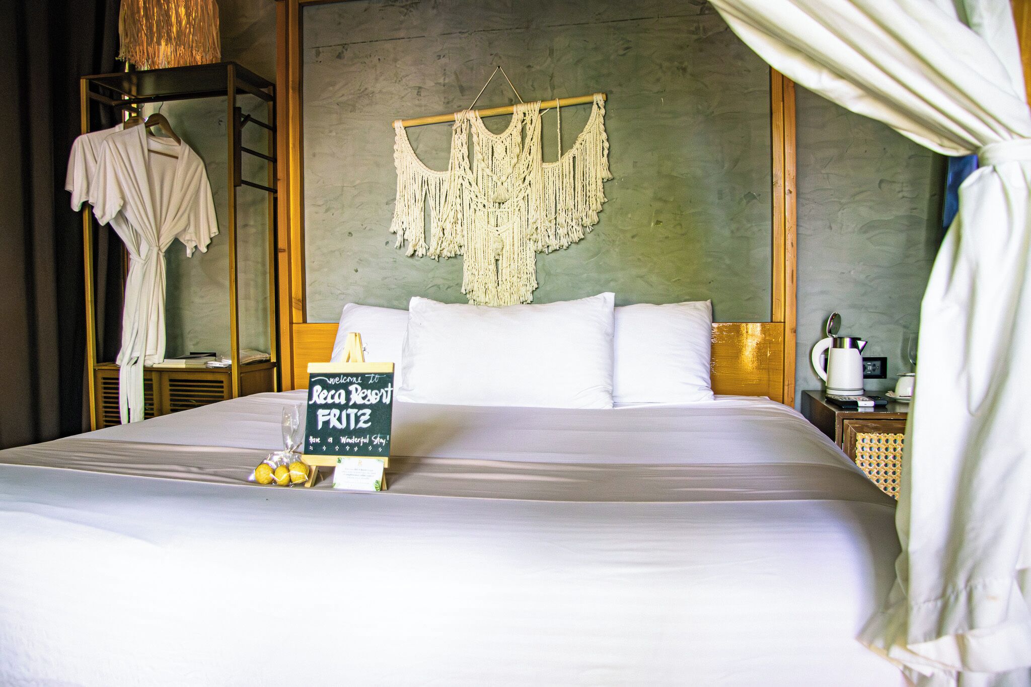 A hotel room with a bed, a welcome sign, a macrame wall hanging, and a robe on a rack.