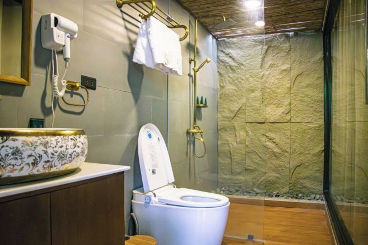 Un baño con inodoro, un lavabo con recipiente decorativo, toallero, cabezal de ducha manual y pared texturizada.