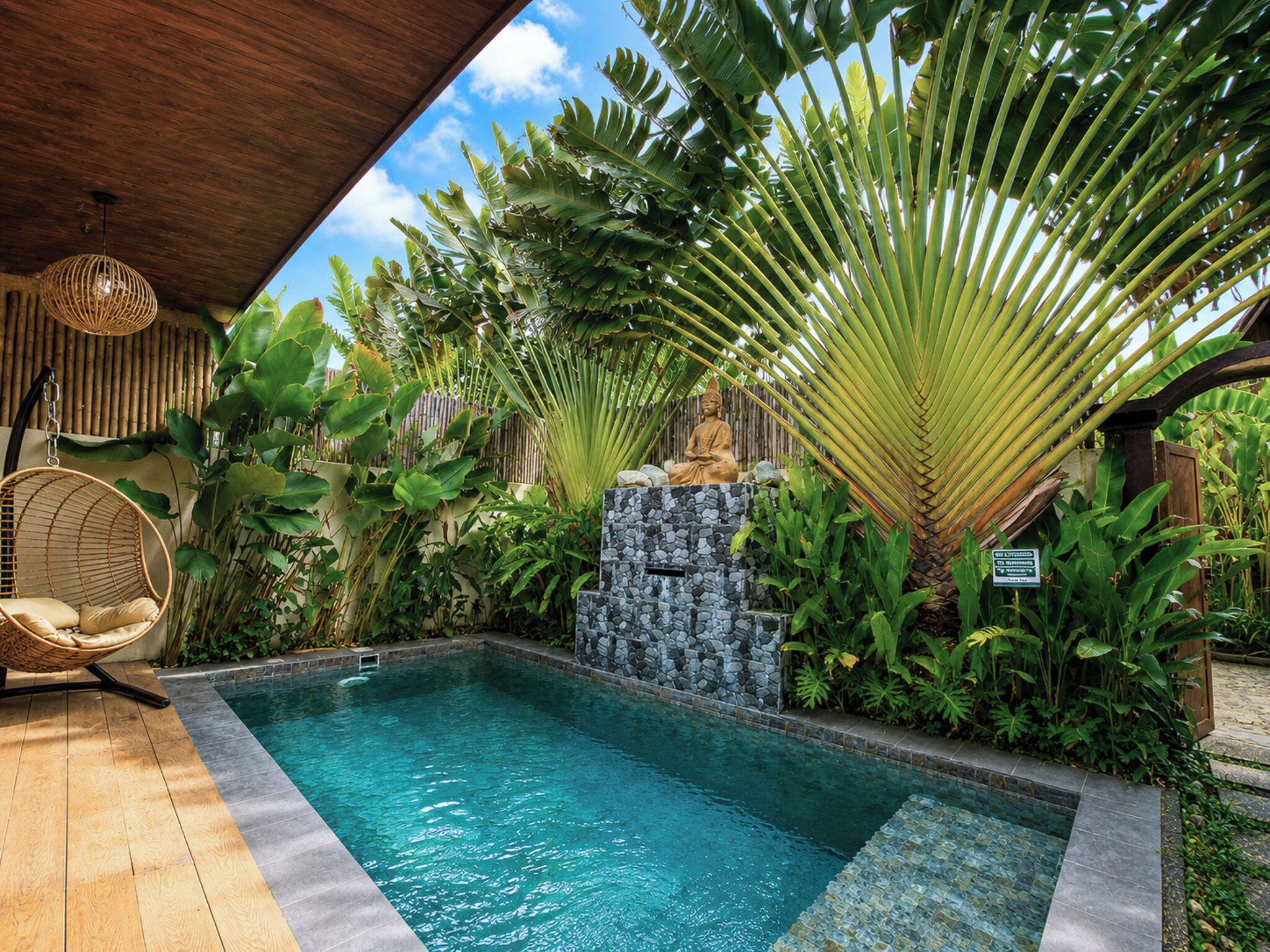 A pool area with a stone waterfall, lush tropical plants, a wooden ceiling, and a wicker chair.