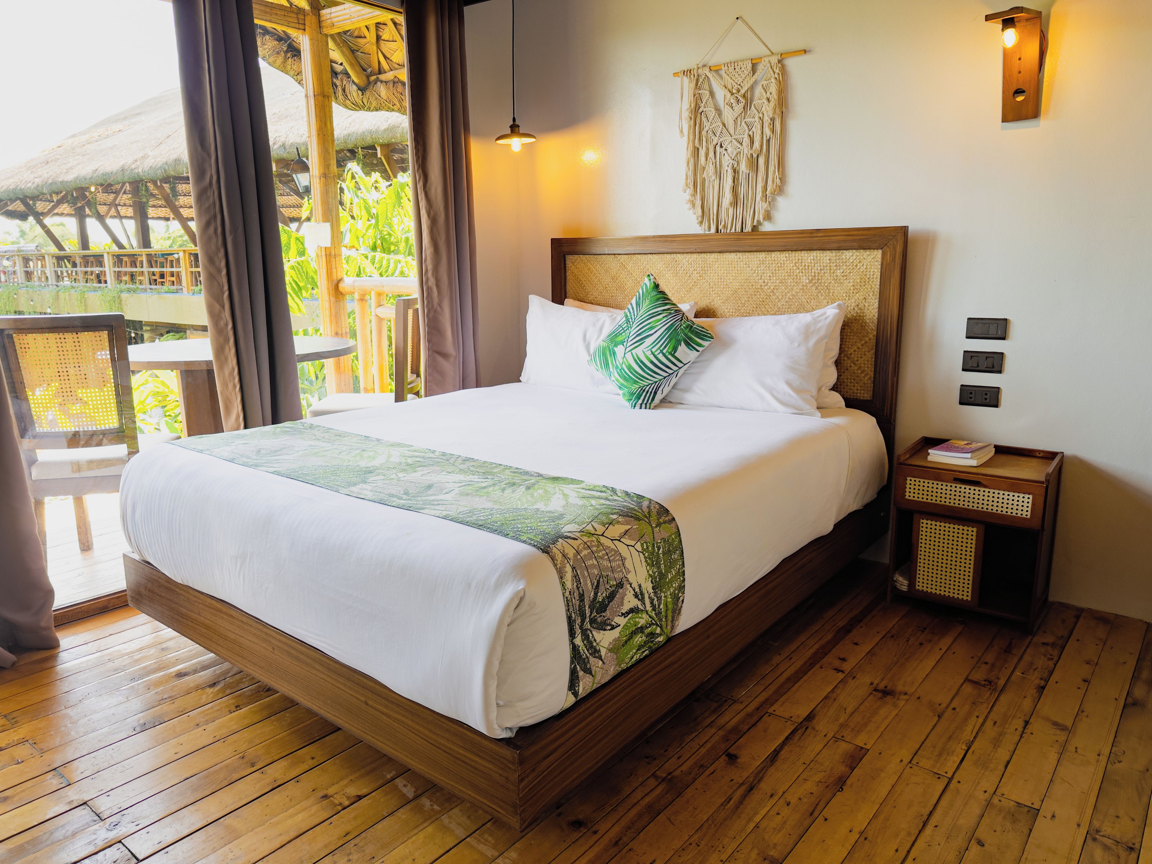 A wooden-framed bed with white bedding, a green leaf-patterned throw, and a macrame wall hanging.