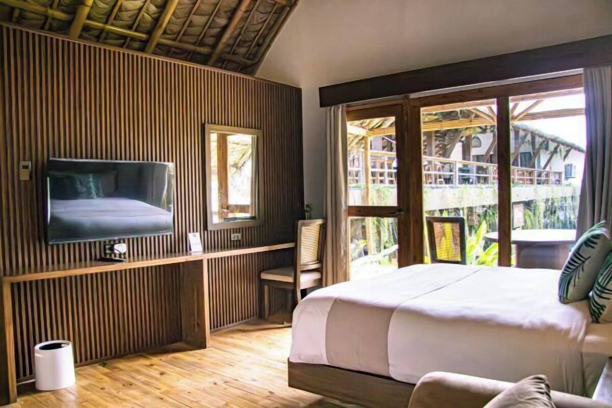 Un dormitorio con cama, televisor, ventana y vista a una estructura de madera en el exterior.
