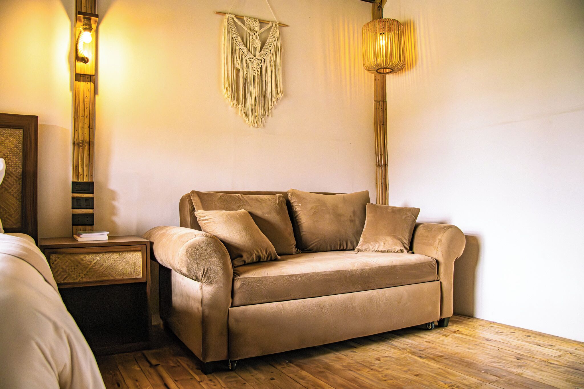 A beige sofa with pillows, a wooden floor, a wall-mounted macrame piece, and a hanging light fixture.
