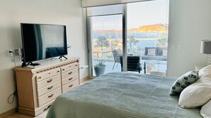 Un dormitorio con cama, televisor sobre una cómoda, y un balcón con vistas al mar y a las montañas.