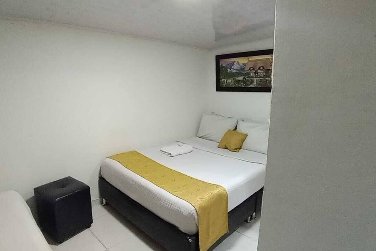 A single-bed room with a white bed, a yellow throw, a black storage box, and a framed picture on the wall.