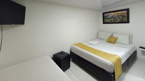 A hotel room with a bed, a small black ottoman, a white bedside table, a flat-screen TV mounted on the wall, and a framed picture above the bed. - NINI nini (Bogotá)