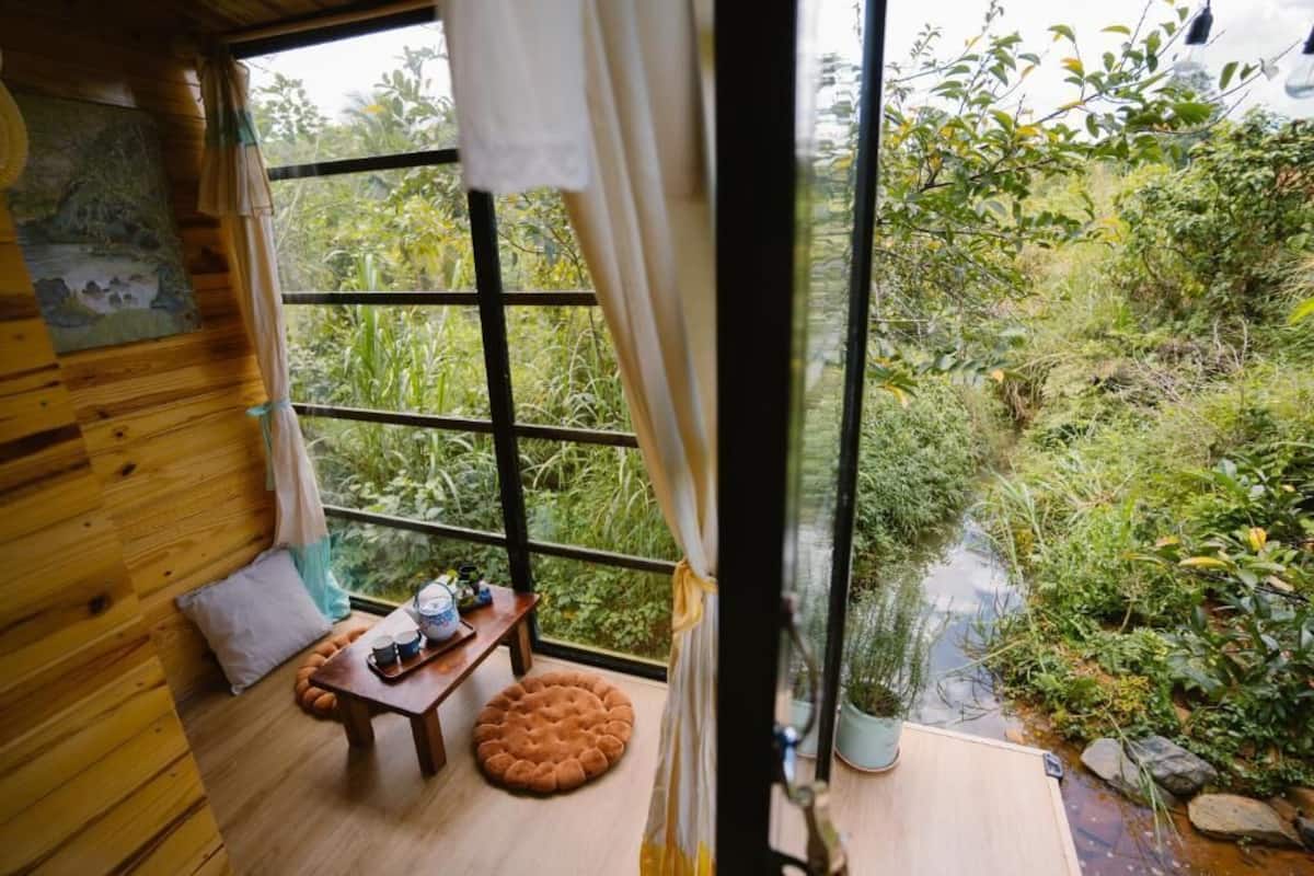 Eine Holzhütte mit einem großen Fenster, das einen üppigen Garten und einen kleinen Teich überblickt.