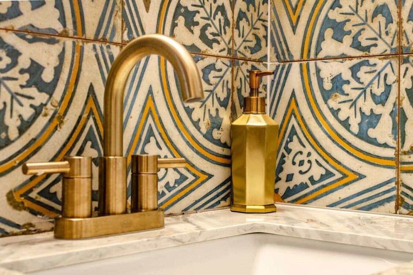 A bathroom with a gold faucet, a gold soap dispenser, and a patterned tile wall.