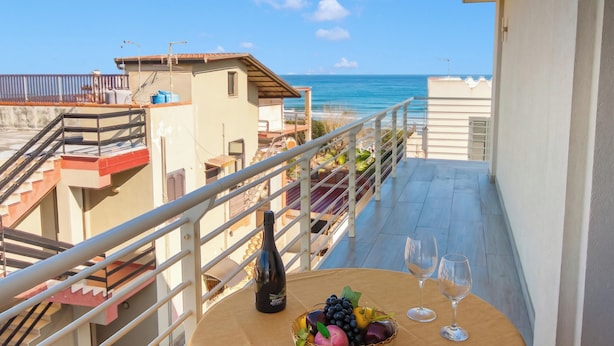 A balcony with a table, a bottle, and glasses, overlooking a beach and buildings.
