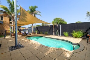 A swimming pool with a curved edge, surrounded by a tiled patio, a shaded area, and a black fence.