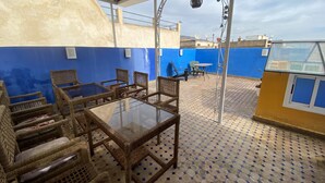 Un espacio al aire libre con techo, amueblado con mobiliario de mimbre y una mesa de vidrio, rodeado de paredes azules y suelo de baldosas.
