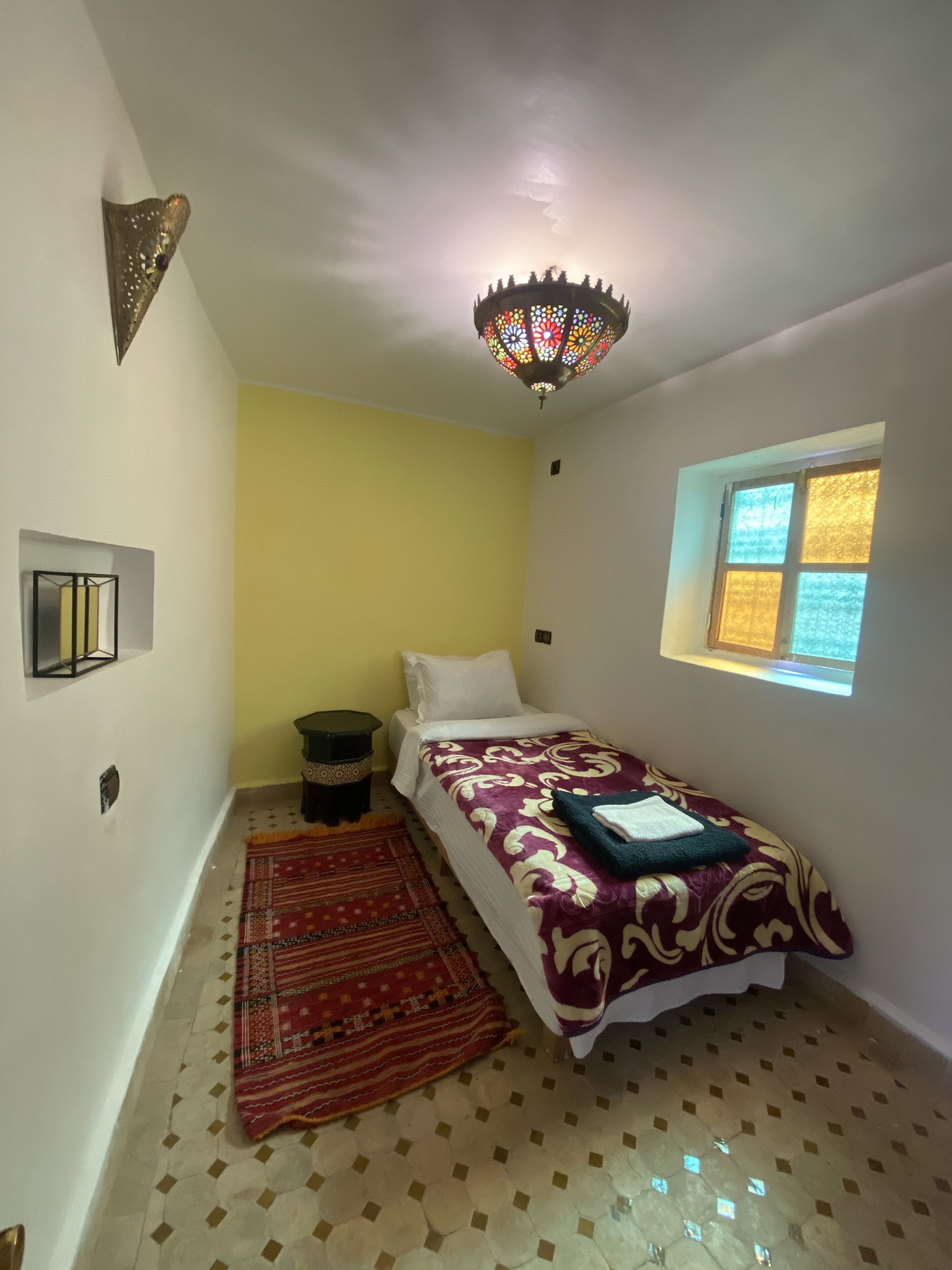 Un dormitorio pequeño y bien iluminado con cama, una lámpara decorativa en el techo, una ventana con vistas y una alfombra estampada.