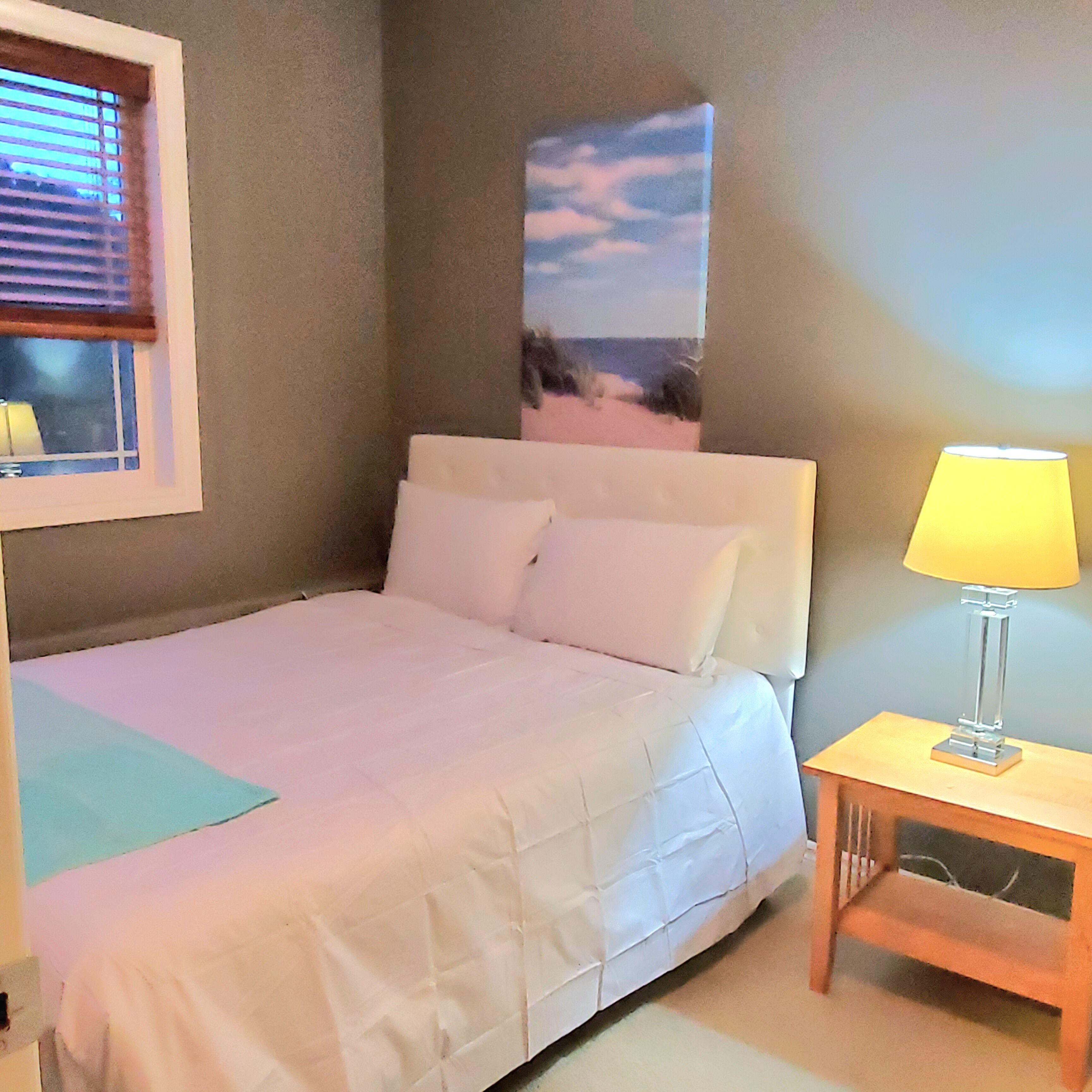A neatly made bed with white linens, a bedside table with a lamp, and a window with blinds.