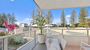 A balcony with a glass table, a potted plant, and a view of a swimming pool and trees. - Ocean and Pool Views from Beachside Apartment! (Gold Coast)
