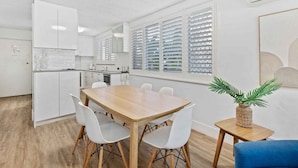 A modern kitchen with white cabinetry, a wooden dining table with white chairs, and a small side table with a plant. - Ocean and Pool Views from Beachside Apartment! (Gold Coast)